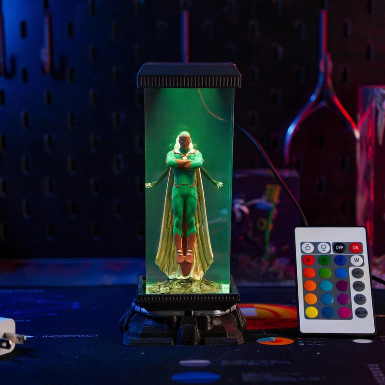 Puede incluir: Figura de acci&oacute;n verde y dorada de un superh&eacute;roe dentro de una vitrina rectangular transparente. La figura tiene los brazos cruzados. La vitrina se asienta sobre una base negra y est&aacute; iluminada con luz verde. Un mando a distancia con opciones de color est&aacute; al lado.
