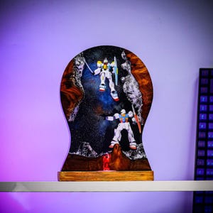 May include: A decorative resin art piece featuring two Gundam model figures in a space-themed setting. The artwork is set within a wooden frame, with a dark background and colorful accents. The figures are in action poses.