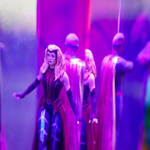 Puede incluir: Figuras de acci&oacute;n, incluyendo un personaje femenino con un traje rojo y negro con cabello rubio, y un personaje masculino con un traje oscuro y capa. Las figuras est&aacute;n sobre un fondo p&uacute;rpura.