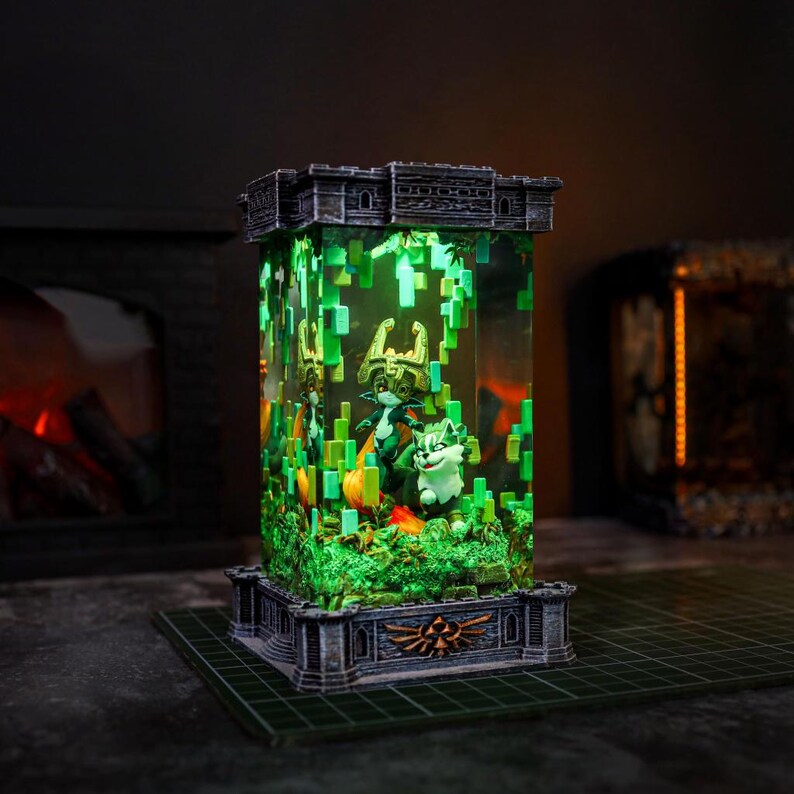 May include: An illuminated display featuring video game characters. The rectangular structure has a castle-like top and base, with a clear central section showcasing figures and green pixelated accents. The base has a gold emblem. The interior is lit with green light.
