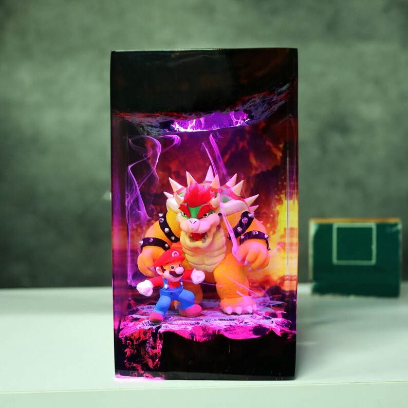 Op de afbeelding: Decoratieve doos met Mario en Bowser uit de Super Mario-videogameserie. Bowser, een groot, oranje schildpadachtig personage met stekels, staat tegenover Mario, een klein figuur in een rode hoed en blauwe overall. De achtergrond heeft een vurige, paarse gloed.