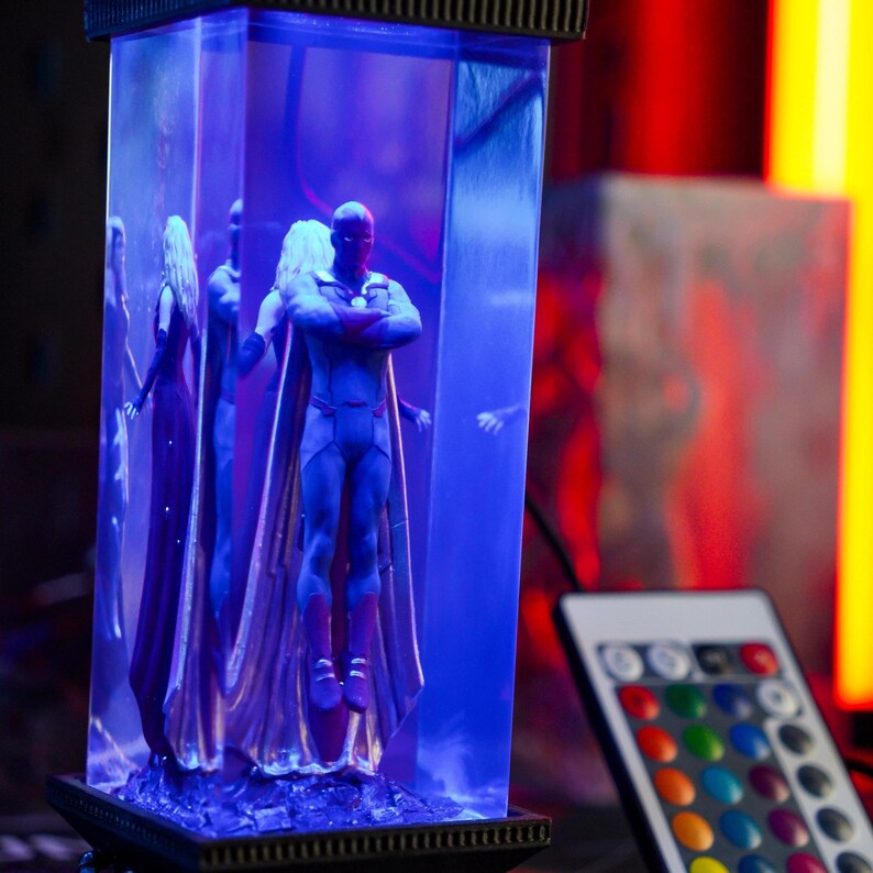 Puede incluir: Una vitrina iluminada en azul con figuras de superh&eacute;roes. La figura central, con un traje azul y rojo, est&aacute; de pie con los brazos cruzados. Otras figuras son visibles dentro de la vitrina. Un mando a distancia con opciones de color est&aacute; en primer plano.