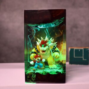 Op de afbeelding: Een harskunstwerk met Mario en Bowser uit de Super Mario-videogame serie. De figuren staan in een rechthoekig blok met een groen gloei-effect. Bowser is geel en Mario draagt zijn kenmerkende rode pet en blauwe overall.