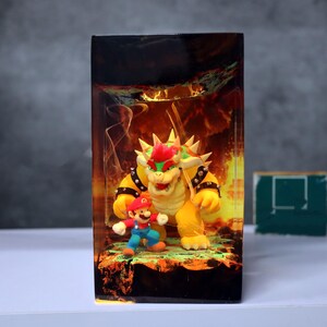 Op de afbeelding: Een rechthoekig kunstwerk van hars met de personages Mario en Bowser uit de Super Mario-videogameserie. Bowser is geel met een rode hanenkam en Mario draagt een rode hoed en een blauwe overall. De achtergrond toont een vurige sc&egrave;ne.