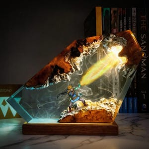 May include: A decorative resin lamp featuring a miniature figure of a character in action, emitting a beam of light. The lamp has a wooden base and a clear, angular resin body with brown and white accents. The scene is illuminated with a warm glow.