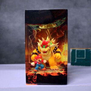 Op de afbeelding: Een rechthoekige display met de personages Mario en Bowser uit de Super Mario-videogame serie. Bowser, een groot geel schildpadachtig wezen met rood haar, staat achter een kleinere Mario-figuur. De achtergrond heeft een vurig, lava-achtig ontwerp.
