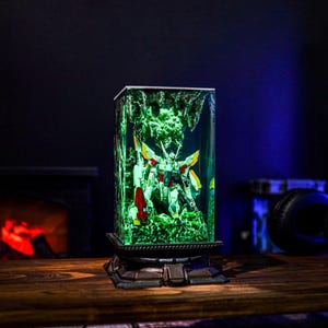 May include: A rectangular, illuminated resin sculpture featuring a detailed robot figure. The sculpture is encased in a clear, rectangular block with a green, glowing interior. The base is a dark, ornate design. The robot has white, yellow, red, and blue accents.