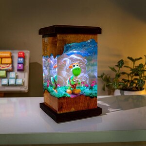 May include: A wooden lamp featuring a green Yoshi figurine in a diorama setting. The lamp has a rectangular shape with a dark brown top and base. The diorama includes a green base, pink mushrooms, and a blue and white background.