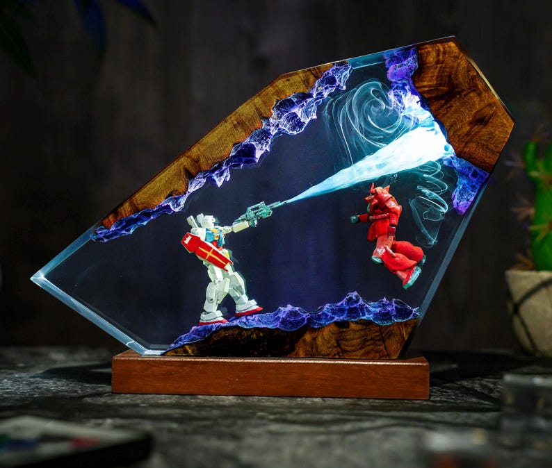 Puede incluir: Una pieza de arte de resina &uacute;nica que presenta una escena de batalla con dos figuras de robots. La obra de arte est&aacute; encerrada en una resina transparente con un marco y una base de madera. Las figuras son blancas, rojas y azules, con un rayo de energ&iacute;a azul. El tema es la ciencia ficci&oacute;n.