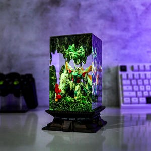 May include: A rectangular, illuminated resin display featuring a detailed Gundam model set within a miniature landscape. The model is predominantly green, white, and yellow, with red accents. The display sits on a black, tiered base.