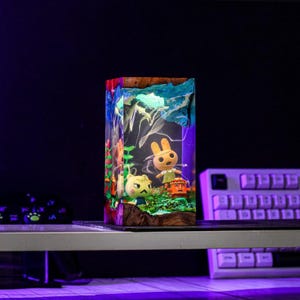 May include: A rectangular resin lamp featuring miniature figurines of characters from the Animal Crossing video game. The lamp has a wooden base and a blue and white top, with colorful details and internal lighting.