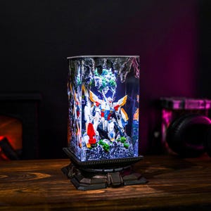 May include: A rectangular, illuminated display featuring a detailed model of a robot. The robot is predominantly white, blue, and orange, set against a backdrop of a miniature landscape. The display sits on a decorative, dark-colored base, with a wooden surface in the foreground.