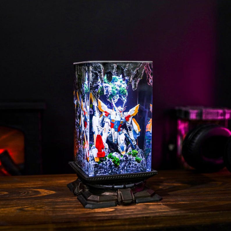 May include: A rectangular, illuminated display featuring a detailed model of a robot. The robot is predominantly white, blue, and orange, set against a backdrop of a miniature landscape. The display sits on a decorative, dark-colored base, with a wooden surface in the foreground.