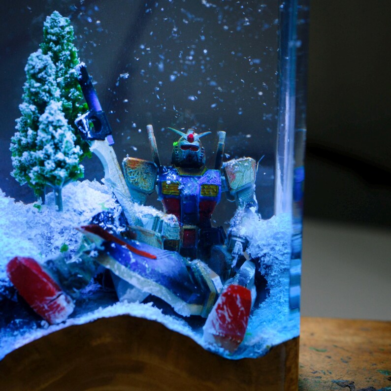 Może przedstawiać: Miniaturowa diorama przedstawiająca kolorową figurę robota na tle zaśnieżonego krajobrazu. Robot jest gł&oacute;wnie niebieski, czerwony i ż&oacute;łty, z precyzyjnym designem. Sztuczny śnieg i miniaturowe drzewa dopełniają zimową scenerię, wszystko w przezroczystej szklanej obudowie.