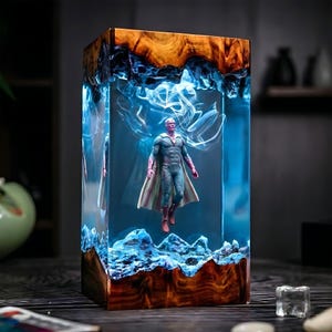 May include: A rectangular resin art piece featuring a superhero figure suspended within a blue-toned, illuminated interior. The base and top showcase natural wood grain, with blue and white swirling patterns. The figure is wearing a cape.