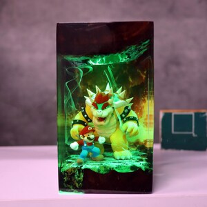 Op de afbeelding: Een harsblok met de personages Mario en Bowser uit de Super Mario-videogame serie. De figuren staan tegen een groen en geel gloeiende achtergrond, met een donkerbruine boven- en onderkant. Het totale effect is een levendige, driedimensionale weergave.