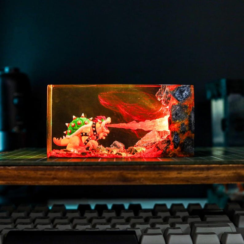 May include: A resin diorama depicting Bowser breathing fire. The scene is set within a rectangular block, featuring a fiery backdrop and rocky landscape. The Bowser figure is detailed with green spikes and a black studded collar.