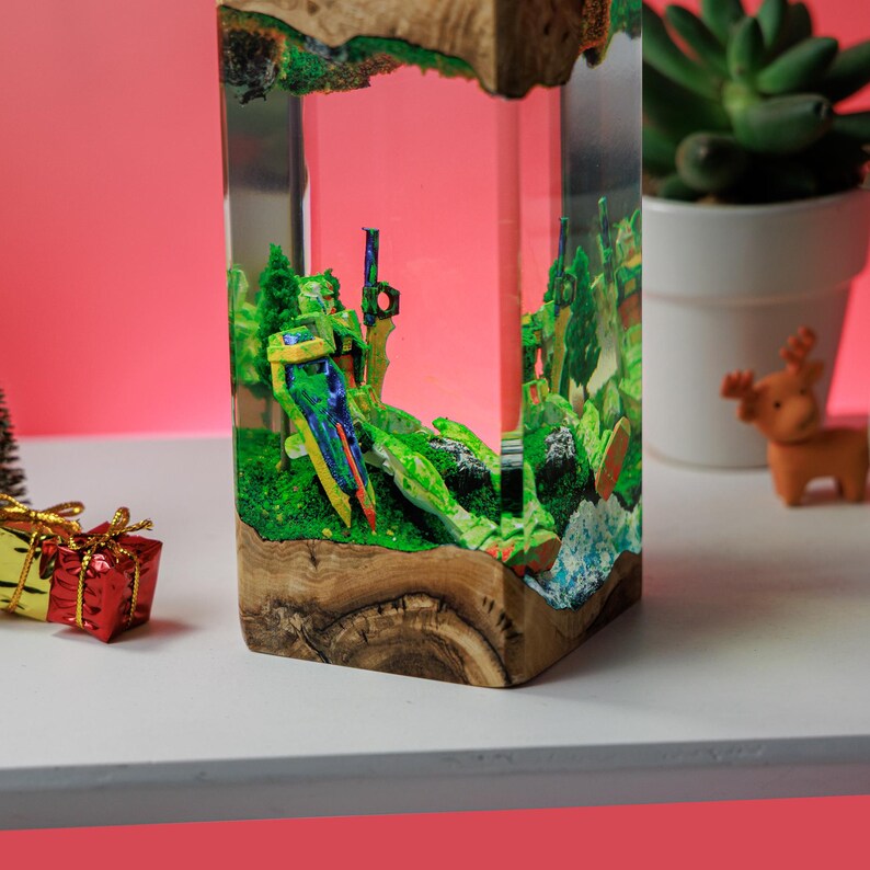 Peut inclure: Une pi&egrave;ce d'art en r&eacute;sine rectangulaire avec une base en bois. La r&eacute;sine transparente renferme un paysage miniature avec un feuillage vert et des &eacute;l&eacute;ments color&eacute;s. Le bord sup&eacute;rieur a une bordure en bois. Un petit coffret cadeau et une plante succulente sont en arri&egrave;re-plan.