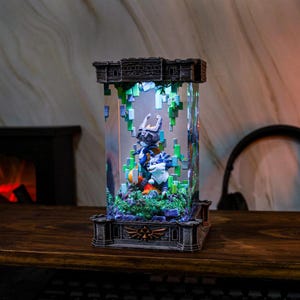 May include: An illuminated display case with a castle-style top and base, housing a detailed diorama of video game characters. The clear central section showcases figures in blue, white, and orange, with green vines and pixelated accents. The case sits on a wooden surface.