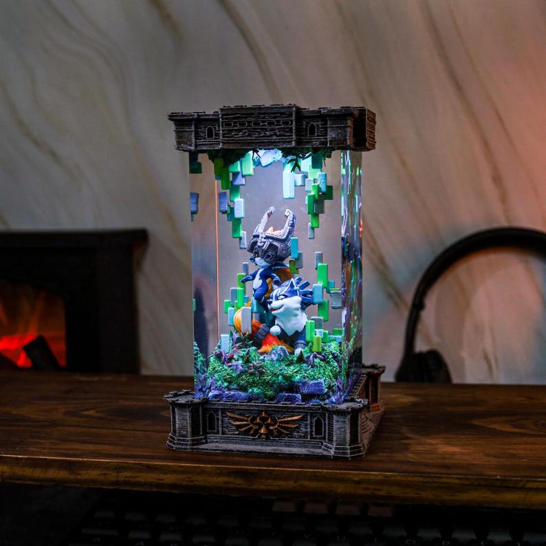 May include: An illuminated display case with a castle-style top and base, housing a detailed diorama of video game characters. The clear central section showcases figures in blue, white, and orange, with green vines and pixelated accents. The case sits on a wooden surface.