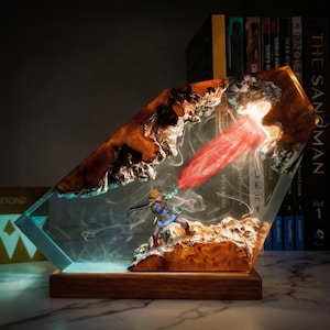 May include: A decorative resin lamp featuring a miniature figure of a character from a video game. The lamp has a wooden base and a clear resin body with embedded wood and a red light effect. The figure is in a dynamic pose, with a red beam emanating from it.