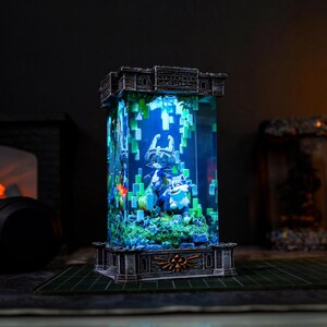 May include: A rectangular, illuminated display with a detailed scene inside, featuring figures and pixelated elements. The display has a stone-like base and top, with a central glass or acrylic enclosure. The interior scene is lit with blue and green hues, creating a vibrant, fantasy-themed aesthetic.