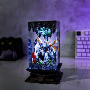 May include: A detailed, illuminated diorama featuring a Gundam model inside a clear, rectangular case. The model is predominantly white, blue, and yellow, set against a backdrop of miniature terrain. The base is dark and ornate.
