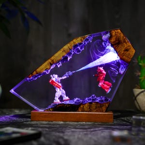Puede incluir: Una pieza de arte en resina &uacute;nica que representa una batalla entre dos figuras de robots. Las figuras est&aacute;n dentro de un bloque de resina transparente, con un marco y base de madera. La escena est&aacute; iluminada con luces LED azules y moradas, creando una exhibici&oacute;n din&aacute;mica.