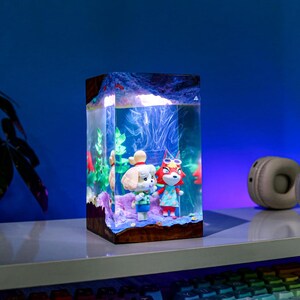 May include: A rectangular resin lamp featuring two Animal Crossing characters inside a clear, illuminated aquarium-like structure. The lamp has a wooden base and a blue and white interior, with red and green accents. A pair of headphones sits on the shelf.