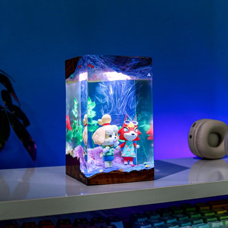 May include: A rectangular resin lamp featuring two Animal Crossing characters inside a clear, illuminated aquarium-like structure. The lamp has a wooden base and a blue and white interior, with red and green accents. A pair of headphones sits on the shelf.