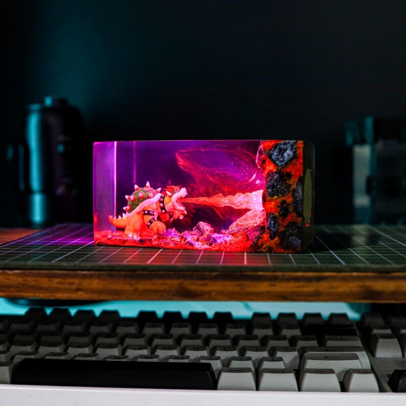 May include: A resin block featuring a Bowser figurine breathing fire. The scene is illuminated with red and purple lighting, with a textured, lava-like edge. The figurine is orange and green.