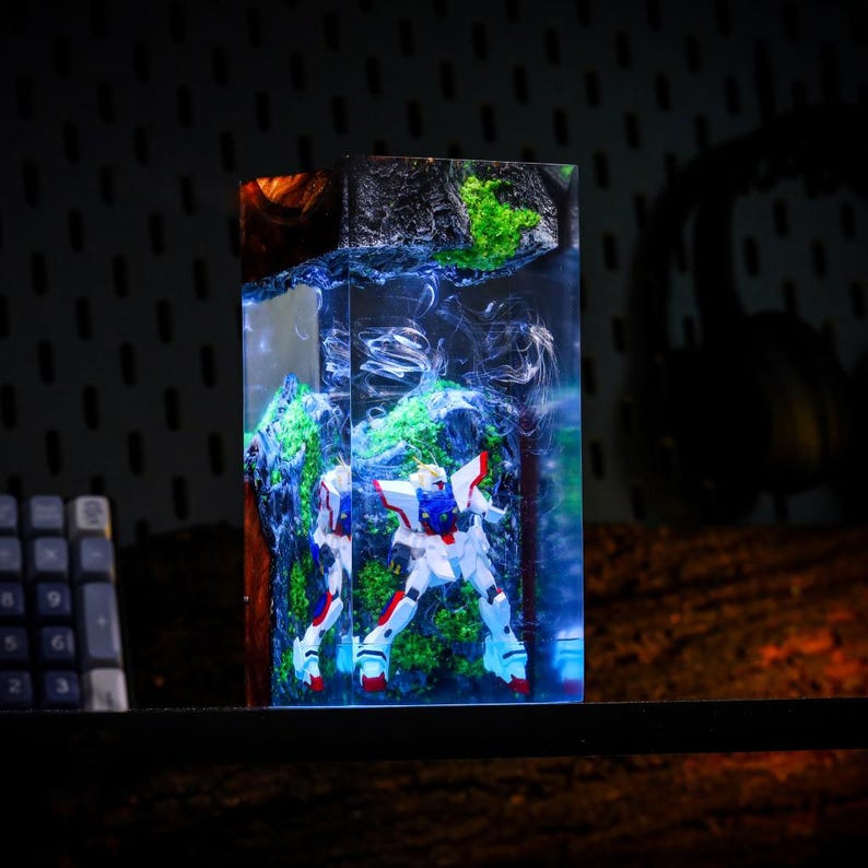 Pu&ograve; includere: Scultura rettangolare in resina con una figura di robot bianco, rosso e blu ambientata in un paesaggio di elementi verdi e blu. La scultura &egrave; racchiusa in resina trasparente, creando un effetto a strati. Lo sfondo &egrave; scuro.