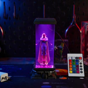 Puede incluir: Una figura de Vision dentro de una vitrina rectangular transparente con base y parte superior negras, iluminada con luz p&uacute;rpura. Un mando a distancia con opciones de color est&aacute; a la derecha.
