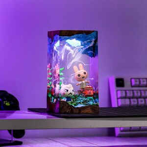 May include: A rectangular resin lamp featuring miniature figurines of cartoon characters. The lamp has a wooden base and a blue and white top, with a light shining through the resin. The figurines include a rabbit and a cat.