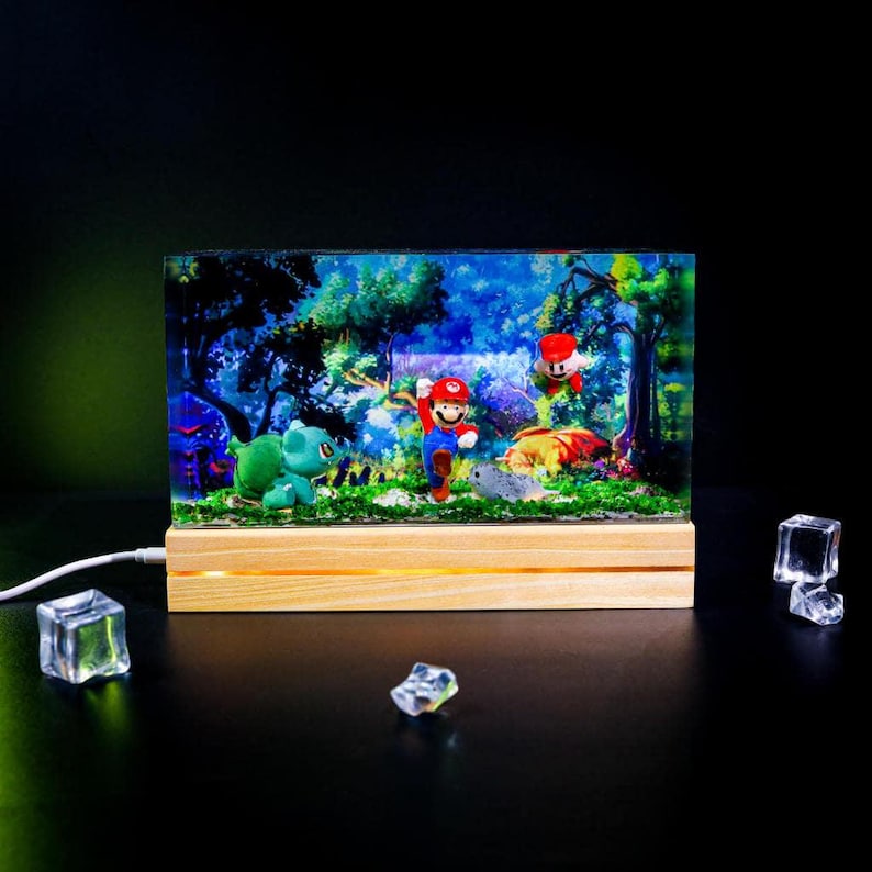 Puede incluir: Una pantalla rectangular iluminada con una escena de bosque con Mario, un Bulbasaur y otros personajes. La pantalla est&aacute; encerrada en material transparente y colocada sobre una base de madera. El fondo representa un bosque azul y verde.