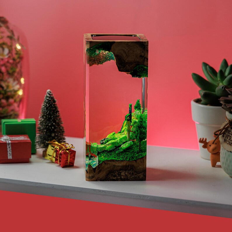 Peut inclure: Une pi&egrave;ce d'art en r&eacute;sine rectangulaire avec une sc&egrave;ne de paysage miniature. La pi&egrave;ce pr&eacute;sente un paysage vert miniature avec des arbres et une cascade, enferm&eacute;s dans de la r&eacute;sine transparente. La base est en bois. Le fond est un d&eacute;grad&eacute; de rouge et de rose.
