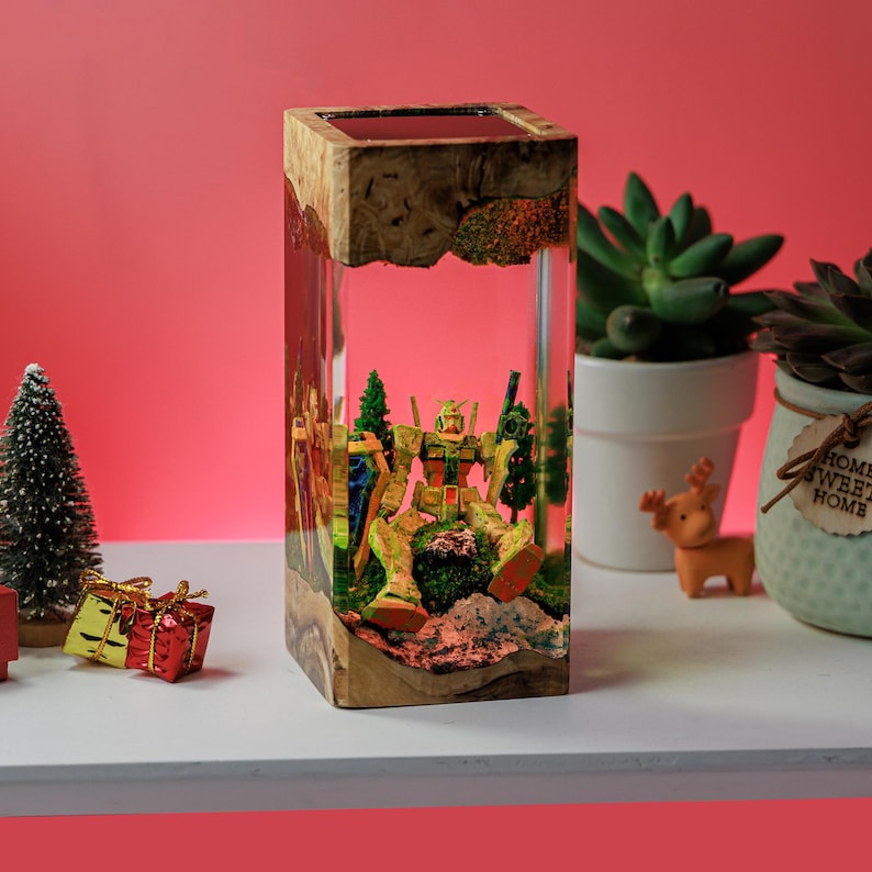 Peut inclure: Une pi&egrave;ce d'art en r&eacute;sine rectangulaire avec une figurine de robot d&eacute;taill&eacute;e, des arbres miniatures et un paysage. La base et le dessus sont en bois. Le fond est rose vif, avec de petits objets d&eacute;coratifs.