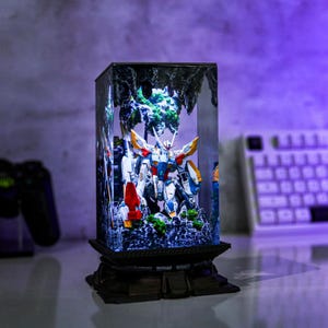 May include: A detailed diorama featuring a Gundam model inside a clear, rectangular display case. The model is predominantly white, blue, red, and yellow, set against a backdrop of miniature terrain and foliage. The case sits on a decorative, dark-colored base.