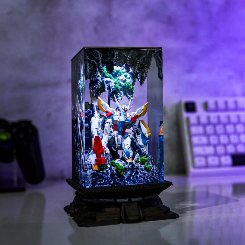 May include: A detailed diorama featuring a Gundam model inside a clear, rectangular display case. The model is predominantly white, blue, red, and yellow, set against a backdrop of miniature terrain and foliage. The case sits on a decorative, dark-colored base.