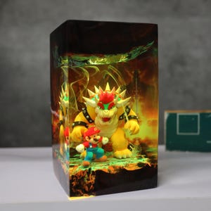 Op de afbeelding: Een rechthoekige harslamp met een sc&egrave;ne met Mario en Bowser uit de Super Mario-videogameserie. De lamp heeft een gele en oranje achtergrond met een groene basis. De personages staan op de voorgrond, met Bowser die boven Mario uittorent.