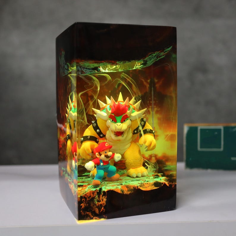 Op de afbeelding: Een rechthoekige harslamp met een sc&egrave;ne met Mario en Bowser uit de Super Mario-videogameserie. De lamp heeft een gele en oranje achtergrond met een groene basis. De personages staan op de voorgrond, met Bowser die boven Mario uittorent.