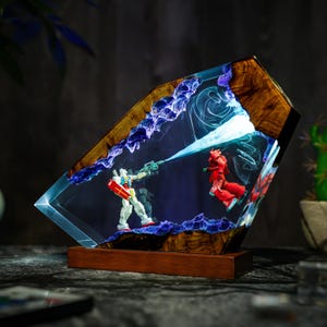 Puede incluir: L&aacute;mpara decorativa de resina con una escena de batalla con dos figuras de robots. La l&aacute;mpara tiene una base y un marco de madera, con detalles en azul y morado dentro de la resina transparente. Un robot dispara un rayo azul al otro.