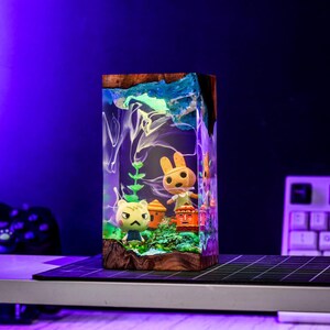 Puede incluir: Una pieza de arte de resina rectangular con figuras en miniatura de personajes del videojuego Animal Crossing. La pieza tiene una base de madera y una parte superior azul y blanca, con los personajes y el escenario encapsulados en la resina transparente.