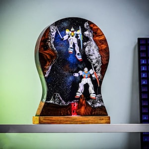 May include: A handcrafted resin sculpture featuring two Gundam robot figures set against a cosmic background. The sculpture has a wooden base and a curved, layered design with brown, white, and blue hues. The robots are white, blue, red, and yellow.