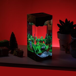 Peut inclure: Lampe rectangulaire en r&eacute;sine avec un diorama de figurines de robots verts dans une for&ecirc;t. La lampe a une base et un dessus noirs, avec les figurines &eacute;clair&eacute;es par une lumi&egrave;re interne. Le fond est rouge vif.
