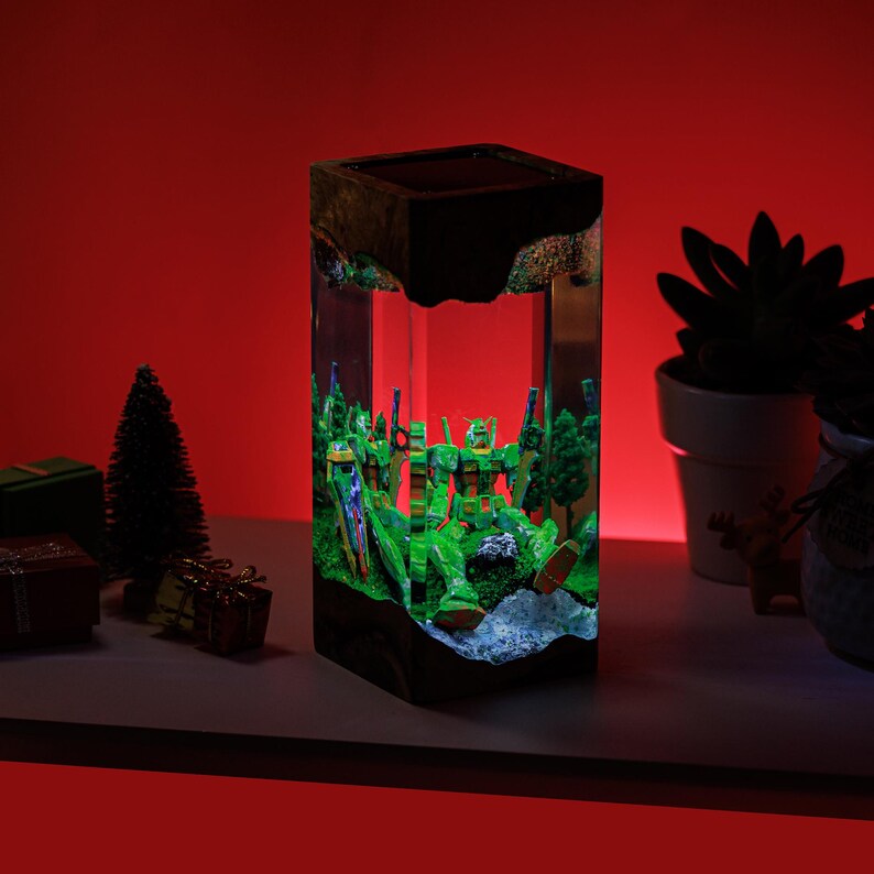 Peut inclure: Lampe rectangulaire en r&eacute;sine avec un diorama de figurines de robots verts dans une for&ecirc;t. La lampe a une base et un dessus noirs, avec les figurines &eacute;clair&eacute;es par une lumi&egrave;re interne. Le fond est rouge vif.