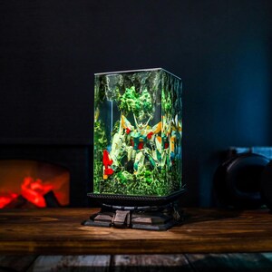 May include: A rectangular lamp featuring a colorful robot figure encased in a clear resin block with green foliage. The lamp sits on a dark, ornate base and is displayed on a wooden surface. The robot has red, yellow, and white accents.