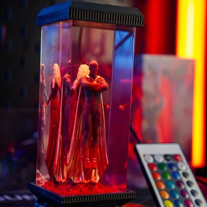 Puede incluir: Una vitrina rectangular de acr&iacute;lico transparente con figuras de personajes con capas rojas y doradas. La vitrina est&aacute; iluminada con luz roja y azul, y tiene una parte superior e inferior negras. Un mando a distancia con opciones de color est&aacute; en primer plano.