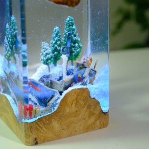 Może przedstawiać: Unikalne dzieło sztuki z żywicy przedstawiające miniaturową dioramę zaśnieżonego krajobrazu z figurką robota i miniaturowymi drzewami. Podstawa wykonana jest z drewna, a scena zamknięta jest w przezroczystej żywicy, tworząc efekt 3D. Robot jest w odcieniach niebieskiego, ż&oacute;łtego i czerwonego.