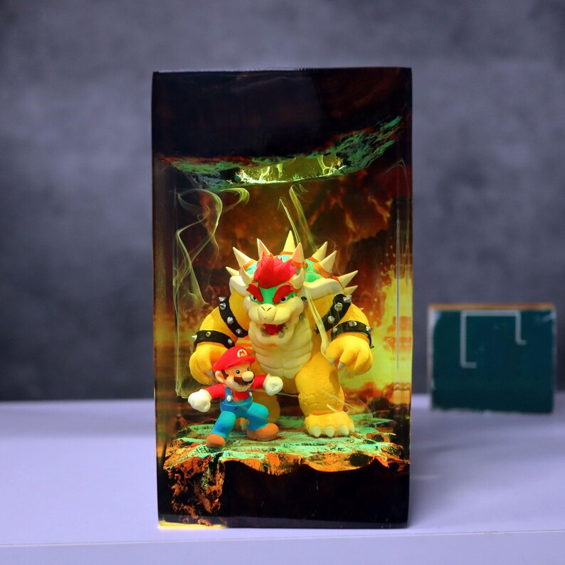 Op de afbeelding: Een harsdiorama met de personages Mario en Bowser uit de Super Mario-videogameserie. Bowser is geel met een rode hanenkam en Mario draagt een rode hoed en een blauwe overall. De achtergrond heeft een vurig, rokerig ontwerp.